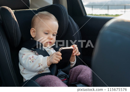 Child is sitting in a car seat, holding a mobile phone in her hands, watching cartoons on the phone Child is sitting in a car seat, holding a mobile phone in her hands, watching cartoons on the phone 115675576