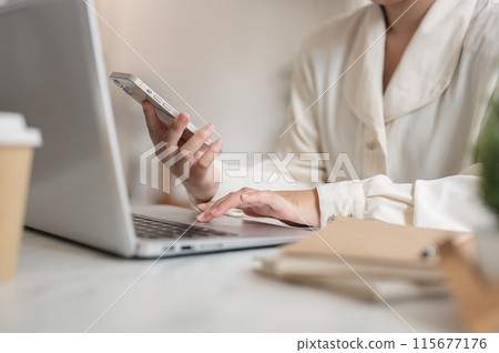 A businesswoman working remotely from a cafe, working on her laptop while using her smartphone. 115677176