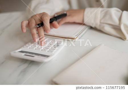 A close-up image of a woman using a calculator at a table, checking and calculating her budget. 115677195