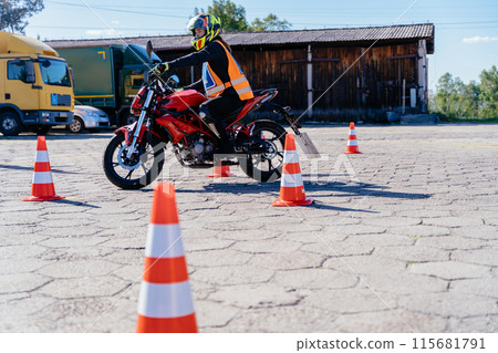 A man is riding a motorcycle in a parking lot with orange and white cones. The cones are arranged in a row, and the man is wearing a helmet 115681791