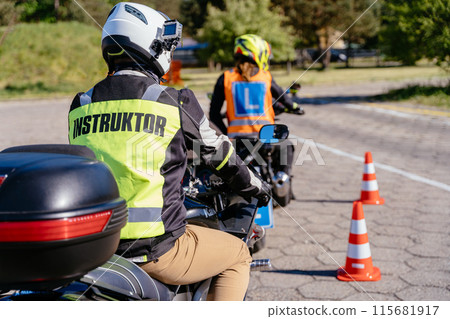 A man in a yellow jacket with the word instructor on it sits on a motorcycle. Another man in a blue vest is riding a motorcycle behind him. Concept of motorcycle school of driving A man in a yellow jacket with the word instructor on it sits on a motorcycle. Another man in a blue vest is riding a motorcycle behind him. Concept of motorcycle school of driving 115681917