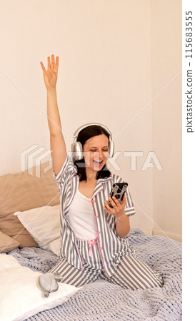 Woman with headphones celebrating, arm raised in bed. 115683555