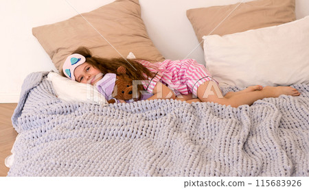 Girl in sleep mask with teddy bear lying on bed. Girl in sleep mask with teddy bear lying on bed. 115683926