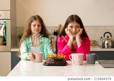 Two girls sitting with birthday cake and mugs at kitchen. Two girls sitting with birthday cake and mugs at kitchen. 115684374
