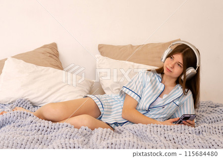 Teenager relaxing in bed with headphones and smartphone, looking thoughtful. 115684480