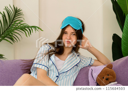 Teenage girl relaxing on couch with sleep mask, smiling gently. 115684503