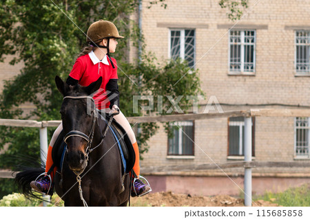 Young rider on horseback looking sideways at stables. 115685858
