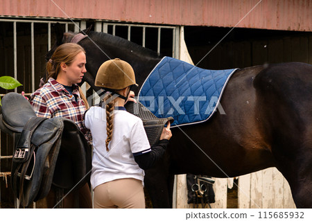 Instructor helping a girl position a saddle on a horse at a stable. 115685932