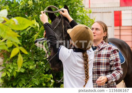 Woman and young rider collaboratively adjusting horse's bridle. Woman and young rider collaboratively adjusting horse's bridle. 115686167
