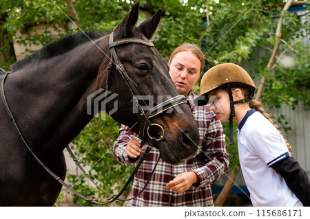 Woman and young rider during bridle adjustment on horse. Woman and young rider during bridle adjustment on horse. 115686171