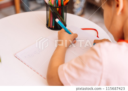 A girl draws with a felt-tip pen with her left hand in a notepad on a white table 115687423