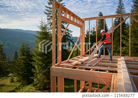 Carpenter constructs wooden-framed house near forest. Man holds large beam in his hands while wearing work clothes and helmet. Idea of modern, eco-friendly construction. Carpenter constructs wooden-framed house near forest. Man holds large beam in his hands while wearing work clothes and helmet. Idea of modern, eco-friendly construction. 115688277