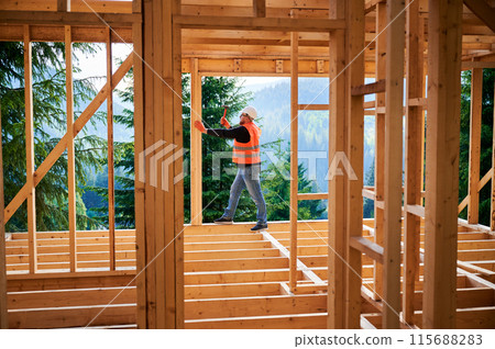 Carpenter constructing two-story wooden frame house near forest. Man hammering nails into the structure, wearing protective helmet and construction vest. Concept of modern ecological construction. Carpenter constructing two-story wooden frame house near forest. Man hammering nails into the structure, wearing protective helmet and construction vest. Concept of modern ecological construction. 115688283
