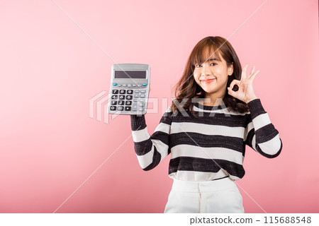 Tax day concept. Woman confident smiling holding electronic calculator and show OK gesture, excited happy Asian female studio shot isolated on pink background, Account and finance counting income Tax day concept. Woman confident smiling holding electronic calculator and show OK gesture, excited happy Asian female studio shot isolated on pink background, Account and finance counting income 115688548