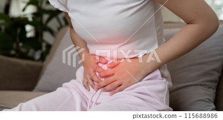 Woman Experiencing Stomach Pain While Sitting on a Couch in a Home Setting, Clutching Her Abdomen in Discomfort 115688986