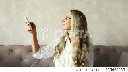 Mature woman turns on the air conditioner using the remote control sitting on couch in room at home 115689635