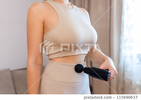 The girl uses a therapeutic percussion vibration massage gun in her hand. Sports recovery concept after training. The girl uses a therapeutic percussion vibration massage gun in her hand. Sports recovery concept after training. 115690637