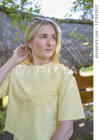 a portrait of a young blond girl in a yellow T-shirt and shorts among the trees on a summer day a portrait of a young blond girl in a yellow T-shirt and shorts among the trees on a summer day 115691622