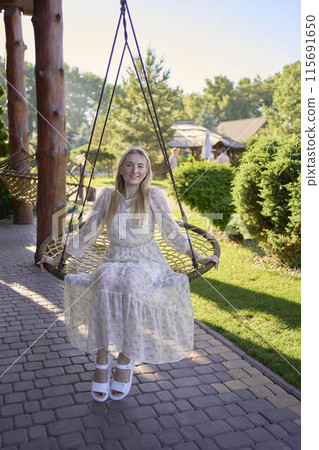 portrait of a young blonde woman in a pastel chiffon dress circling in a wicker hammock 115691650