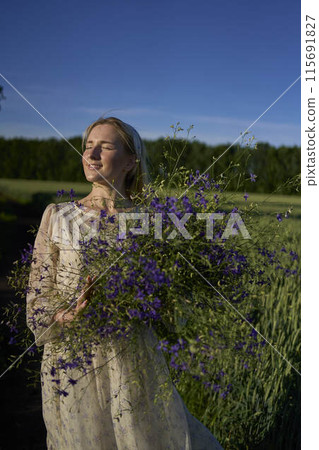 portrait of a young blonde woman in a pastel chiffon dress walking with a giant bouquet of wild flowers in a field 115691827
