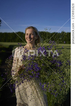 portrait of a young blonde woman in a pastel chiffon dress walking with a giant bouquet of wild flowers in a field 115691828