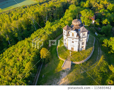 Aerial view of the Church of Saint John the Baptist and Our Lady of Mount Carmel surrounded by lush greenery at Makova Hora, Czechia, on a bright spring day. 115693462