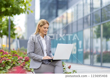 A young woman in a gray suit is sitting on a bench near an office building, holding a laptop on her lap and typing on the keyboard. 115695408