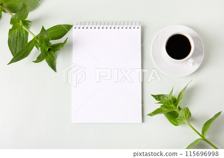 Composition with opened blank notepad, cup of black coffee, green leaves of zinnia on a gray background. Festive office desktop concept. Morning coffee cup. 115696998