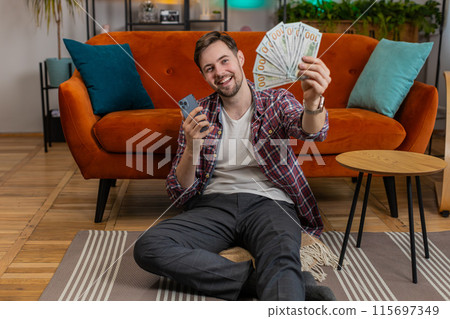Successful happy rich man holds smartphone waving dollar bill money fan sitting in room at home Successful happy rich man holds smartphone waving dollar bill money fan sitting in room at home 115697349