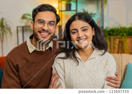 Loving happy smiling young Indian couple spending leisure time sitting arm around relaxing on sofa 115697350