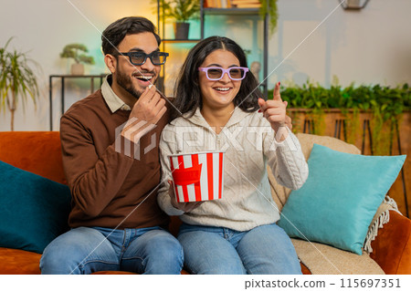 Cheerful young Indian couple in 3D glasses eating popcorn and watching comedy movie sitting on sofa 115697351