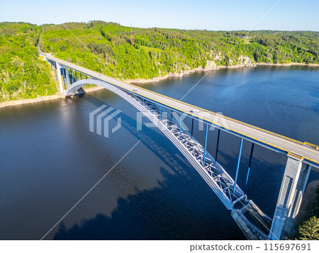 An aerial view of the Zdakov Bridge crossing the Vltava River, surrounded by lush green trees. An aerial view of the Zdakov Bridge crossing the Vltava River, surrounded by lush green trees. 115697691