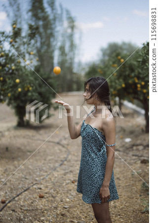 A woman in a summer dress floats an orange in an orchard, embodying a leisurely outdoor activity A woman in a summer dress floats an orange in an orchard, embodying a leisurely outdoor activity 115698831