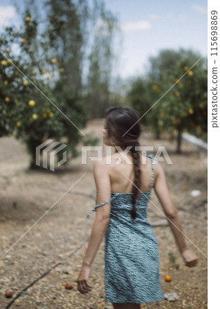 A stylish woman with long braid dressed in a blue dress is seen walking in an orchard 115698869