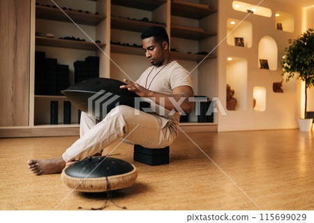 Low-angle view of African young man in trance sitting playing on steel tongue drum by hands on floor at home with cozy interior. Silence calm filling room by sounds of music from musical instrument. Low-angle view of African young man in trance sitting playing on steel tongue drum by hands on floor at home with cozy interior. Silence calm filling room by sounds of music from musical instrument. 115699029