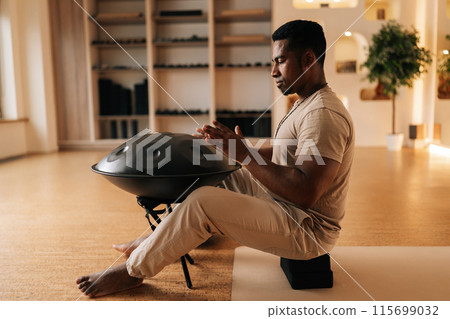 Side view of meditated black male in trance practicing native music traditional drumming instrument playing with hands sitting on floor in lotus pose. Relaxed African man drumming rhythm on tank drum Side view of meditated black male in trance practicing native music traditional drumming instrument playing with hands sitting on floor in lotus pose. Relaxed African man drumming rhythm on tank drum 115699032