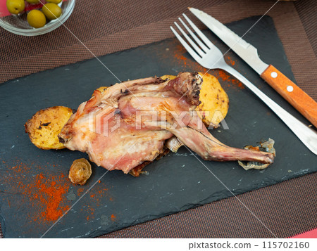 Baked half of rabbit carcass with several slices of potatoes is served on black board Baked half of rabbit carcass with several slices of potatoes is served on black board 115702160