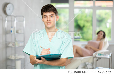 Smiling man beautician in uniform standing in cosmetology clinic, filling out clipboard with medical records Smiling man beautician in uniform standing in cosmetology clinic, filling out clipboard with medical records 115702676