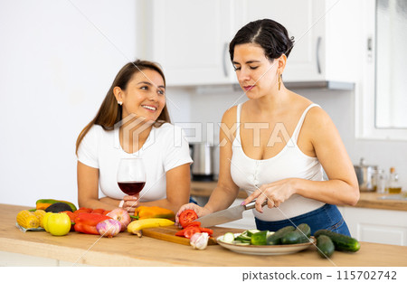 Two happy female friends preparing salad and drinking red wine together in kitchen Two happy female friends preparing salad and drinking red wine together in kitchen 115702742