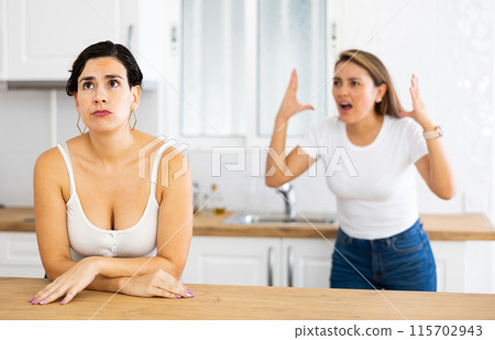 Frustrated young woman listening to angry girlfriend in home kitchen Frustrated young woman listening to angry girlfriend in home kitchen 115702943