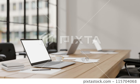 A laptop computer with a white screen mockup on a meeting table in a modern meeting room. 115704068