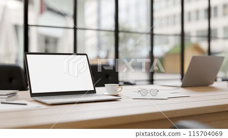A laptop computer with a white screen mockup on a meeting table in a modern meeting room. 115704069