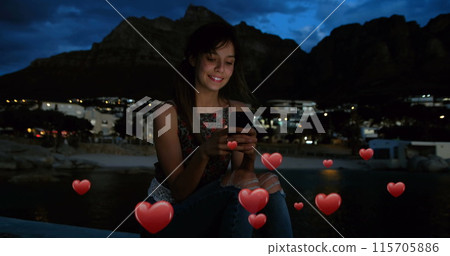 Digital composite of a Caucasian woman sitting on an edge near the beach at night and digital hearts Digital composite of a Caucasian woman sitting on an edge near the beach at night and digital hearts 115705886