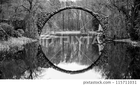 Rakotz Bridge, German: Rakotzbrucke, forms a perfect stone arch reflected in the calm waters of a serene German lake, surrounded by bare trees. Black and white image. 115711031