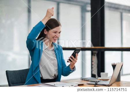 A young businesswoman in a blue blazer celebrates success while looking at her smartphone in a modern office setting. 115714751
