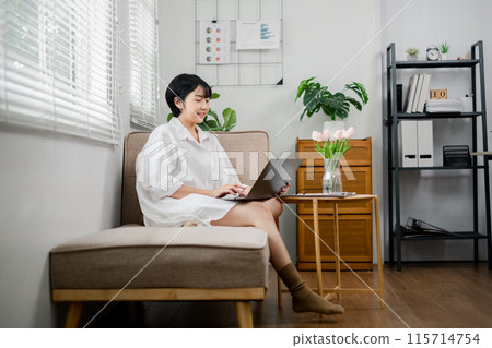 A woman is sitting on a couch in a living room, working on her laptop. She is smiling and she is enjoying her work. The room is decorated with potted plants and a vase of flowers, giving it a cozy 115714754