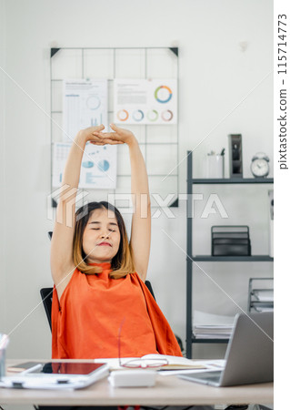 Young woman in orange top stretching at her office desk, surrounded by modern workspace elements including charts and shelves. 115714773