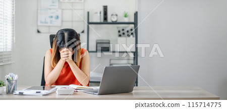 A woman is sitting at a desk with a laptop and a stack of papers. She is looking at the laptop and she is in a state of distress A woman is sitting at a desk with a laptop and a stack of papers. She is looking at the laptop and she is in a state of distress 115714775