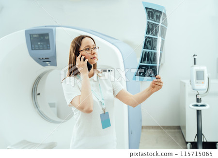 Medical female specialist holding x-ray of patient's brain and looking at it. Confident Caucasian doctor consults patient over the phone inspecting roentgen in front of of MRI scanner. 115715065