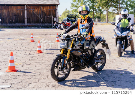 Motorcycle driving school concept. Man in yellow vest is riding a motorcycle in a parking lot. Several other motorcycles in lot, and a few people are standing around. Scene is casual and relaxed. Motorcycle driving school concept. Man in yellow vest is riding a motorcycle in a parking lot. Several other motorcycles in lot, and a few people are standing around. Scene is casual and relaxed. 115715294